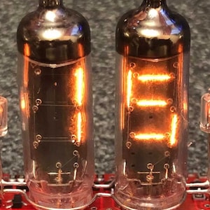 May include: Two clear glass Nixie tubes with glowing orange filaments forming the numbers "11" on a red circuit board with black resistors.