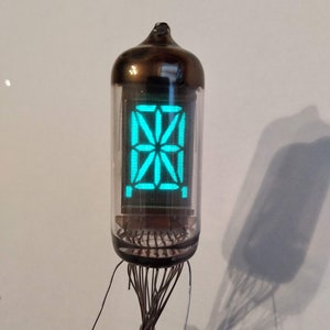 May include: A vintage Nixie tube display with a green glow. The tube is in a glass enclosure with a metal top and bottom. The display shows a pattern of lines and dots.