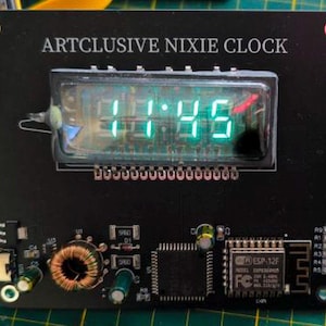 Puede incluir: Una placa de circuito rectangular negra etiquetada como ARTCLUSIVE NIXIE CLOCK con una pantalla digital central que muestra números verdes iluminados. La placa presenta componentes electrónicos expuestos, incluyendo una bobina, chips y un puerto USB.