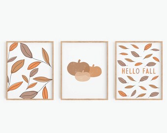 May include: Three framed prints with fall-themed designs. The first print features a pattern of orange and brown leaves. The second print features three pumpkins in shades of orange and brown. The third print features a pattern of orange and brown leaves with the text "HELLO FALL" in orange.
