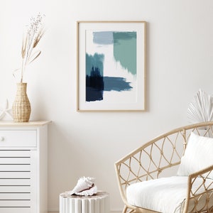 May include: A framed abstract painting with blue and green brushstrokes on a white background. The painting is hanging on a white wall above a white dresser and a wicker chair with a white cushion.