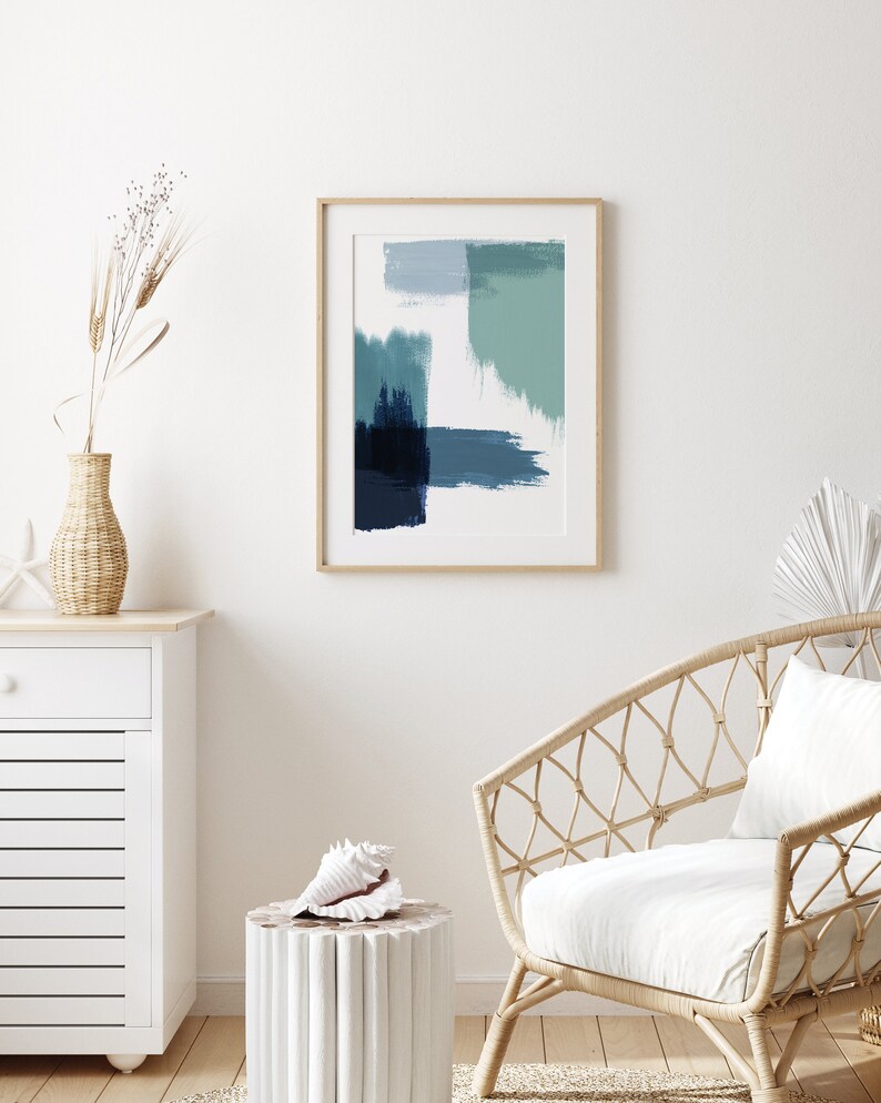 May include: A framed abstract painting with blue and green brushstrokes on a white background. The painting is hanging on a white wall above a white dresser and a wicker chair with a white cushion.