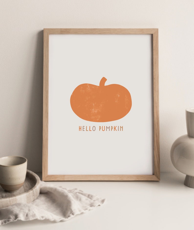Może przedstawiać: Wydruk w ramie z prostym pomarańczowym sylwetem dyni i tekstem "HELLO PUMPKIN" w kolorze białym.