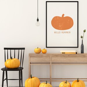 Może przedstawiać: Czarny obraz w ramie z pomarańczową ilustracją dyni i tekstem "HELLO PUMPKIN". Obraz wisi na białej ścianie nad drewnianym stolikiem konsolowym z kilkoma pomarańczowymi dyniami na g&oacute;rze.