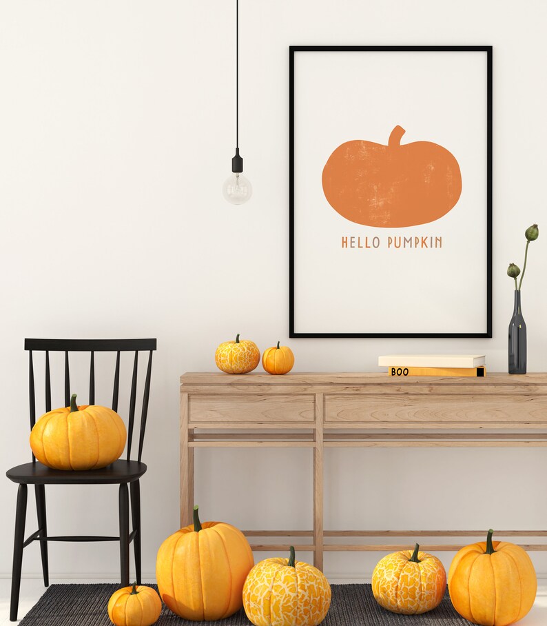 Może przedstawiać: Czarny obraz w ramie z pomarańczową ilustracją dyni i tekstem "HELLO PUMPKIN". Obraz wisi na białej ścianie nad drewnianym stolikiem konsolowym z kilkoma pomarańczowymi dyniami na g&oacute;rze.
