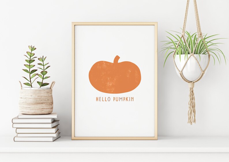 Może przedstawiać: Wydruk w ramie z prostym pomarańczowym sylwetowym obrazem dyni i tekstem "HELLO PUMPKIN". Wydruk jest na białym tle i jest oprawiony w jasną drewnianą ramę.
