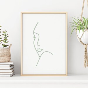 K&ouml;nnte beinhalten: Ein gerahmter Druck mit einer minimalistischen Linienzeichnung eines Frauen Gesichts in Salbeigr&uuml;n. Der Druck ist auf wei&szlig;em Hintergrund und in einem hellen Holzrahmen gerahmt. Der Druck steht auf einem wei&szlig;en Regal mit einer Topfpflanze auf jeder Seite.