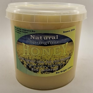 May include: A clear plastic bucket filled with light-colored honey, featuring a label that reads Natural Springtime HONEY, From rapeseed, willow, fruit trees blossoms, Collected in Lithuania, Raw, unfiltered, Season 2026, Net Weight 1500 g.