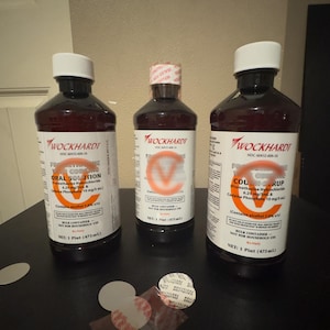 May include: Three brown bottles of Wockhardt oral solution and cough syrup with white labels and caps. The labels include text and orange circular designs. The bottles are on a dark surface.