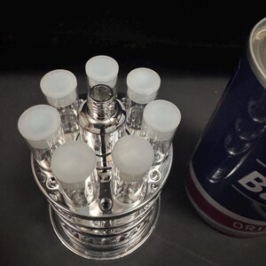 May include: A silver shot glass set with six clear glasses, each with a frosted white cap, arranged around a central chrome liquor dispenser. A blue and white can is partially visible in the background.