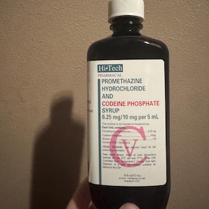 May include: A white ceramic mug with a black handle and the text "But First, Coffee" in a bold, black font. The mug is set against a blurred background, suggesting a cozy setting.