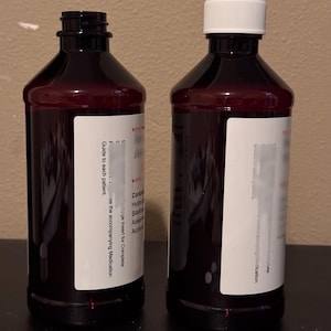 May include: Two dark red plastic bottles with white labels. One bottle has a white cap, the other is open. The labels have text, including "Guide to each patient" and "ge Insert for Complete". The bottles are on a dark surface.