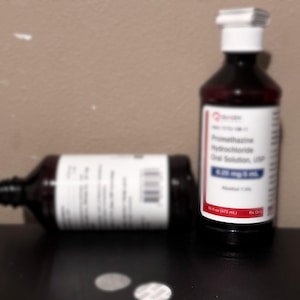 May include: Two dark brown medicine bottles with white labels and red accents. One bottle is upright, the other is lying on its side. The labels read "Promethazine Hydrochloride Oral Solution, USP 6.25 mg/5 ml Alcohol 7.0% 16 fl oz (473 mL)."