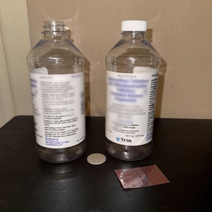 May include: Two clear plastic bottles with white labels and caps. One bottle is empty, the other contains a liquid suspension. The labels include text and measurements of 16 fl oz (473 ml). A small, round, silver disc and a red foil packet are also present.