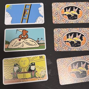 May include: A collection of tarot cards featuring whimsical illustrations. One card shows a bird climbing a ladder, another a bear in a dress, and a third with two frogs and a sun. The backs of the cards have a decorative pattern.