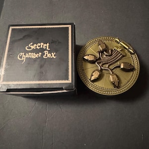 May include: A square black box with the text Secret Chamber Box printed in gold, accompanied by a round, metallic gold-toned container featuring a decorative lid adorned with five sculpted, snake-like figures connected by thin, textured metal chains.