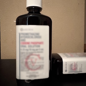 May include: A black bottle with a white label and a white cap. The label has blurred text, including the name "AKORN". Another bottle is lying on its side. The bottles contain a liquid oral solution.