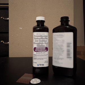 May include: Two dark brown medicine bottles with white labels and caps. One bottle has a white label with the text "Alcohol 8% (v/v)" and "16 fl oz (473 mL)". The other bottle is empty. The bottles are on a dark surface.