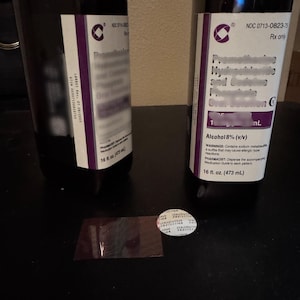 May include: Two dark bottles with white and purple labels, containing a liquid. The labels include text and a logo. One bottle has a visible measurement of 16 fl. oz. (473 mL). A clear seal and a small, rectangular piece of plastic are on the surface.