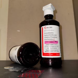 May include: Two dark brown medicine bottles, one upright and one on its side. The upright bottle has a white label with red and black text, including the brand "QUAGEN" and the volume, 16 fl oz (473 mL). The other bottle is empty.
