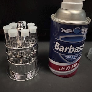 May include: A chrome shot glass holder with clear shot glasses and a can of Barbasol shaving cream. The shaving cream can is blue with white text and a red stripe. The shot glass holder has two tiers and is made of metal.