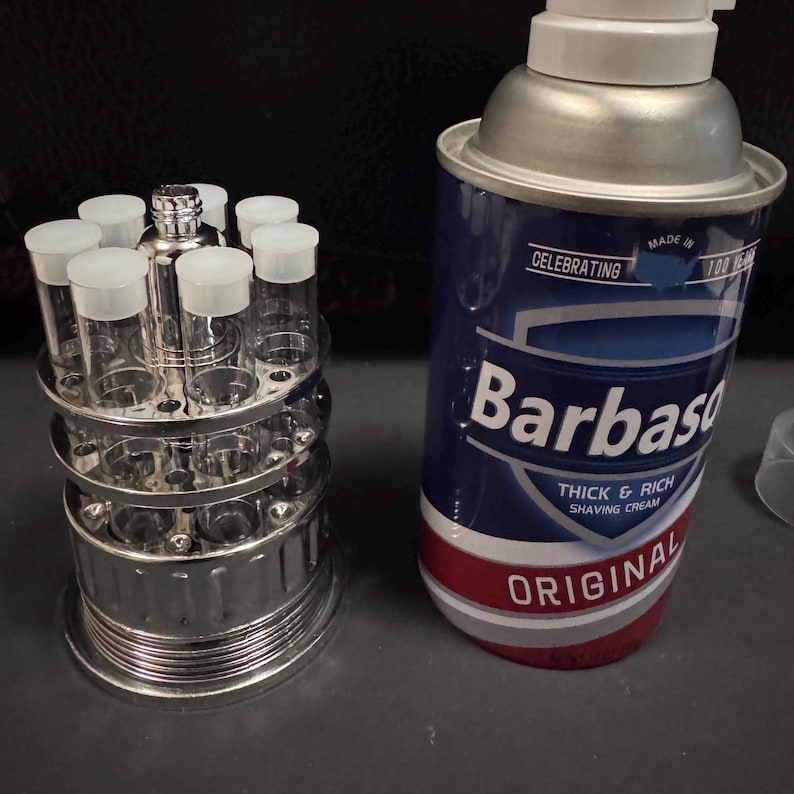 May include: A chrome shot glass holder with clear shot glasses and a can of Barbasol shaving cream. The shaving cream can is blue with white text and a red stripe. The shot glass holder has two tiers and is made of metal.