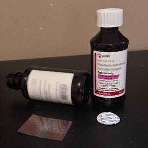 K&ouml;nnte beinhalten: Zwei dunkelbraune Medizinflaschen mit wei&szlig;en Etiketten und Verschl&uuml;ssen. Eine Flasche steht aufrecht, die andere liegt auf der Seite. Auf den Etiketten steht "Oral Solution" und enthalten Dosierungsinformationen. Ein Foliensiegel und ein Flaschensiegel sind ebenfalls vorhanden.