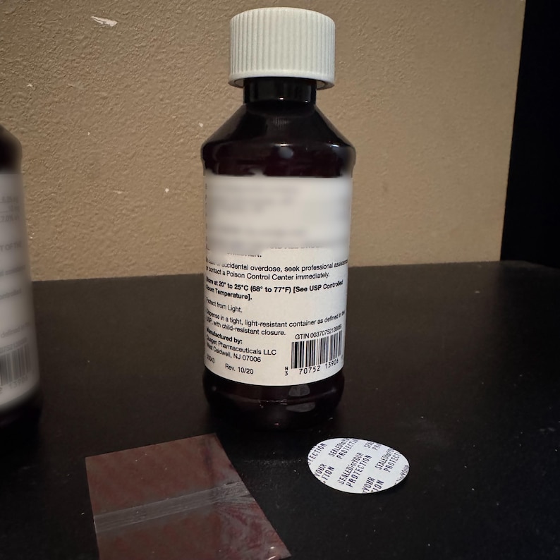 K&ouml;nnte beinhalten: Eine braune Flasche mit wei&szlig;em Etikett und wei&szlig;em Verschluss. Die Flasche steht auf einer schwarzen Oberfl&auml;che. Ein kleiner, klarer Plastikbeutel und ein wei&szlig;er Siegel mit Text befinden sich ebenfalls auf der Oberfl&auml;che. Die Flasche enth&auml;lt Fl&uuml;ssigkeit.