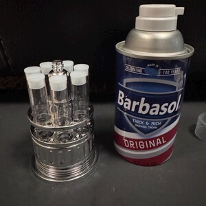 May include: A can of Barbasol shaving cream with a blue, white, and red label, celebrating 100 years. Beside it is a chrome stand holding clear glass vials with white caps. The shaving cream can is silver and white.