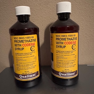 May include: Two bottles of Promethazine with Codeine Syrup, each with a white cap and a yellow label. The label includes the brand name Qualitest and the text "ONE PINT (473 ml)". The bottles are filled with a dark liquid.