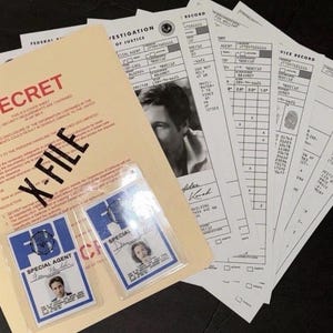 May include: A collection of FBI-themed props, including a manila folder labeled "SECRET" and "X-FILE", along with several official-looking documents. Two FBI Special Agent ID cards are visible. The documents feature text and a black and white photo.