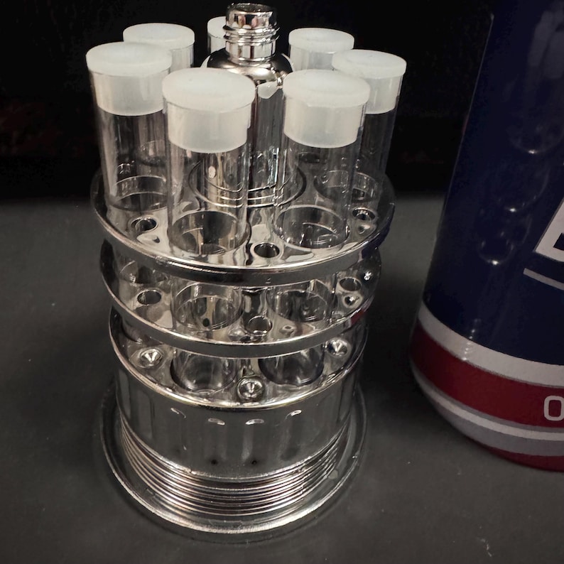 May include: A shiny chrome test tube rack holding clear glass tubes with white caps. The rack has multiple levels and circular holes for the tubes. A cylindrical container with a blue and red design is in the background.