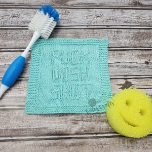 Peut inclure: Un torchon à vaisselle en tricot bleu sarcelle avec le texte "F*ck the Dishes" en blanc. Le torchon est sur une surface en bois avec une brosse à bouteille bleue et blanche et une éponge jaune.