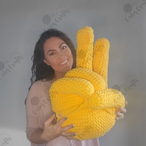 Pu&ograve; includere: Una grande mano gialla all'uncinetto che fa il segno della pace. La mano &egrave; tenuta da una persona che indossa una camicia rosa.