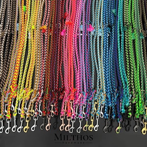 Pode incluir: Uma coleção de coleiras para cães trançadas coloridas com fechos de metal prateado e preto. As coleiras estão dispostas em filas e apresentam uma variedade de cores brilhantes, incluindo rosa, azul, verde, amarelo e laranja. As coleiras são feitas à mão e apresentam o texto "MILTHOS - handmade" na parte inferior da imagem.