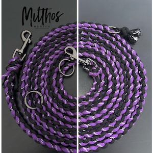 Puede incluir: Una correa para perro trenzada negra y morada con detalles plateados y dos clips metálicos. La correa está enrollada y tiene un pequeño borla negra trenzada en un extremo.
