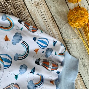 Puede incluir: Tela blanca con un patr&oacute;n de globos aerost&aacute;ticos azules, naranjas y marrones. La tela tiene un ribete azul alrededor de los bordes.