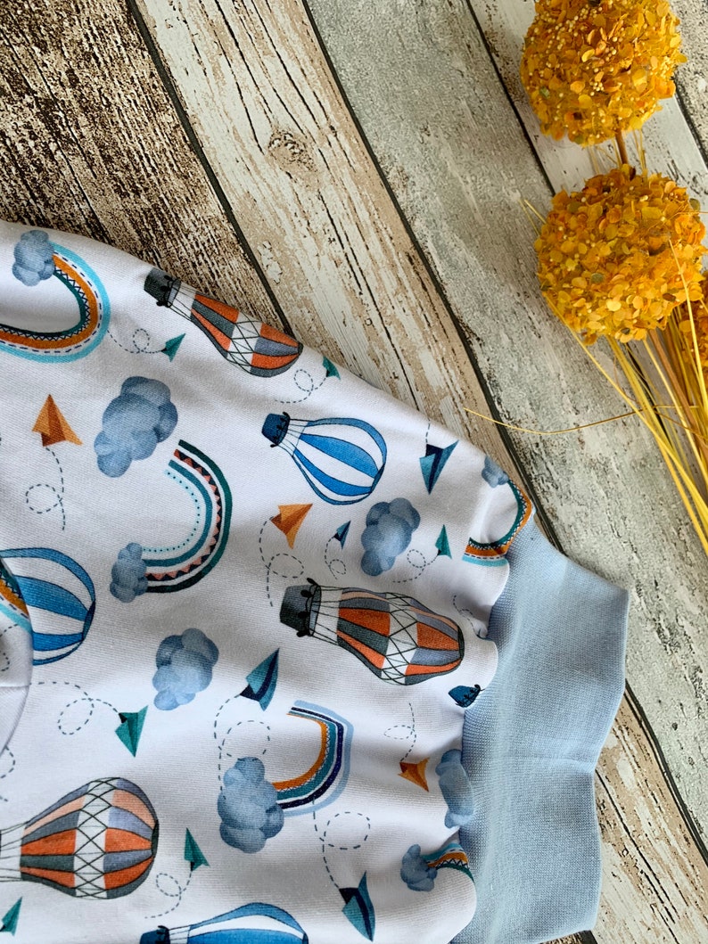 Puede incluir: Tela blanca con un patr&oacute;n de globos aerost&aacute;ticos azules, naranjas y marrones. La tela tiene un ribete azul alrededor de los bordes.