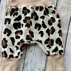 Op de afbeelding: Babybroek met een beige tailleband en manchetten. De broek heeft een witte stof met een zwart en mauve luipaardprintpatroon. De stof lijkt zacht en getextureerd.