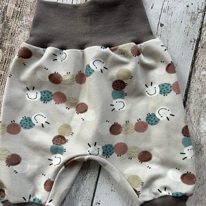 Könnte beinhalten: Babyhose mit hellbeigem Hintergrund und einem Muster aus bunten Kreisen und lächelnden Gesichtern. Der Bund und die Bündchen sind einfarbig dunkelgrau. Die Hose ist aus weichem Stoff.
