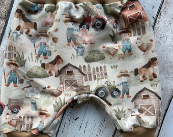 Puede incluir: Pantalones de bebé con estampado de granja. Los pantalones tienen una cintura y puños marrones, y una tela beige con ilustraciones de animales de granja, graneros, tractores y granjeros. El diseño incluye detalles en marrón, rojo, verde y azul.