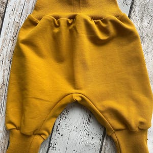 以下が含まれることがあります： マスタードイエローのベビーパンツ。幅広のゴムウエストバンドとカフ付きの脚が特徴です。