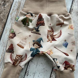 May include: Beige baby pants with a whimsical print featuring owls, wizards, spell books, and potion bottles. The pants have a high, tan waistband and matching cuffs. The overall design is a playful, cartoon-like illustration.