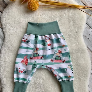Könnte beinhalten: Babyhose mit einem skurrilen Zwerg- und Weihnachtsbaum-Muster. Die Hose hat einen grünen Bund und Bündchen mit einem weiß-grünen Streifenhintergrund. Die Zwerge tragen rote Hüte und halten Geschenke. Die Hose ist aus weichem Stoff.