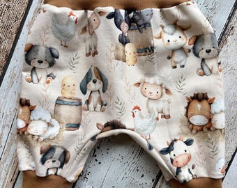 Puede incluir: Pantalones de tela color crema con un estampado de acuarela repetitivo de varios animales de granja, incluyendo vacas, ovejas, caballos, pollos y perros. La prenda tiene una cintura ancha de color marrón sólido y puños de tobillo a juego.