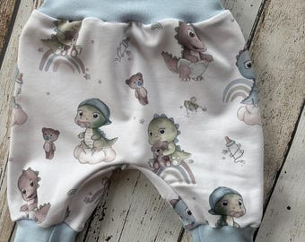 Puede incluir: Pantalones de bebé con cintura y puños azul claro. La tela blanca está estampada con dibujos de dinosaurios, arcoíris, ositos de peluche y biberones en colores pastel. Los pantalones tienen un corte holgado.