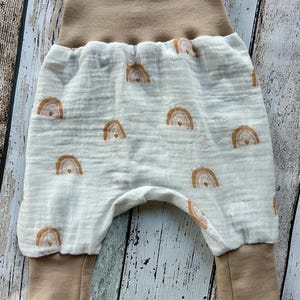 May include: Baby pants with a white background and a pattern of brown and tan rainbows. The waistband and cuffs are tan. The pants are made of a soft, textured fabric.