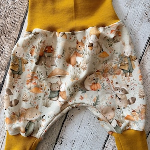 Könnte beinhalten: Babyhose aus cremefarbenem Stoff mit einem wiederkehrenden Muster aus Waldtieren wie Kaninchen, Eichhörnchen, Igeln und Waschbären zwischen Pilzen und Herbstblättern, abgerundet mit gerippten Bündchen und einem Taillenbund in Senfgelb.