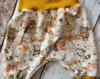 Puede incluir: Pantalones para bebé con tela color crema y un patrón repetitivo de animales del bosque, incluyendo conejos, ardillas, erizos y mapaches entre setas y hojas de otoño, con puños y cintura acanalados en color amarillo mostaza sólido.