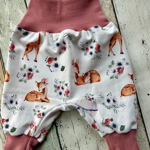 Può includere: Pantaloni da bambino con sfondo bianco, con un motivo di cervi e composizioni floreali nei toni del marrone, rosa e blu. La vita e i polsini sono di colore rosa antico. I pantaloni hanno una vestibilità ampia.